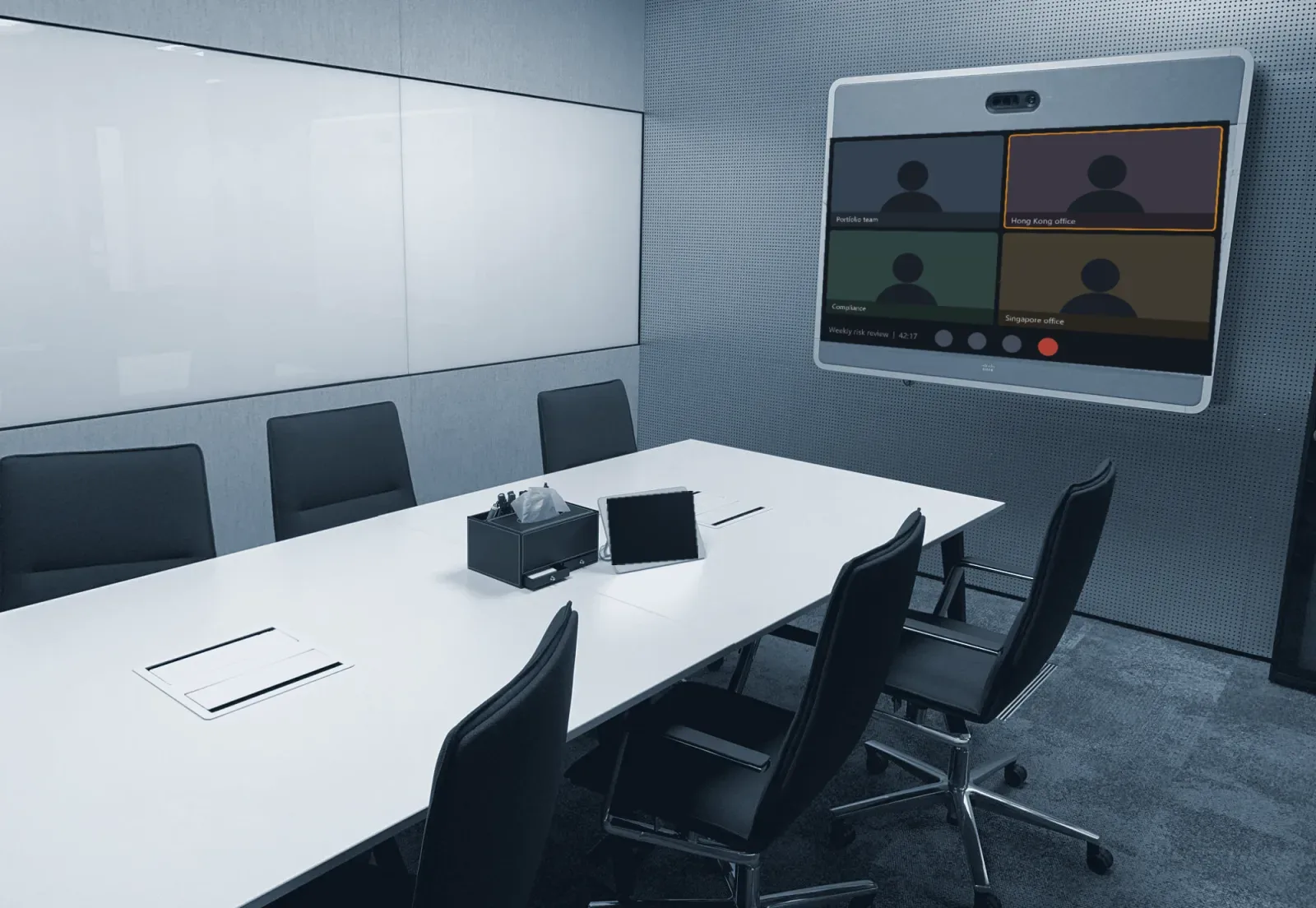 A meeting room with a wall-mounted video conferencing screen and a touch panel on the table